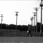 A still from the film Berman's March where a man walks from the right of the frame to the right along a road, holding a jug, with electrical lines in the distance beside him.